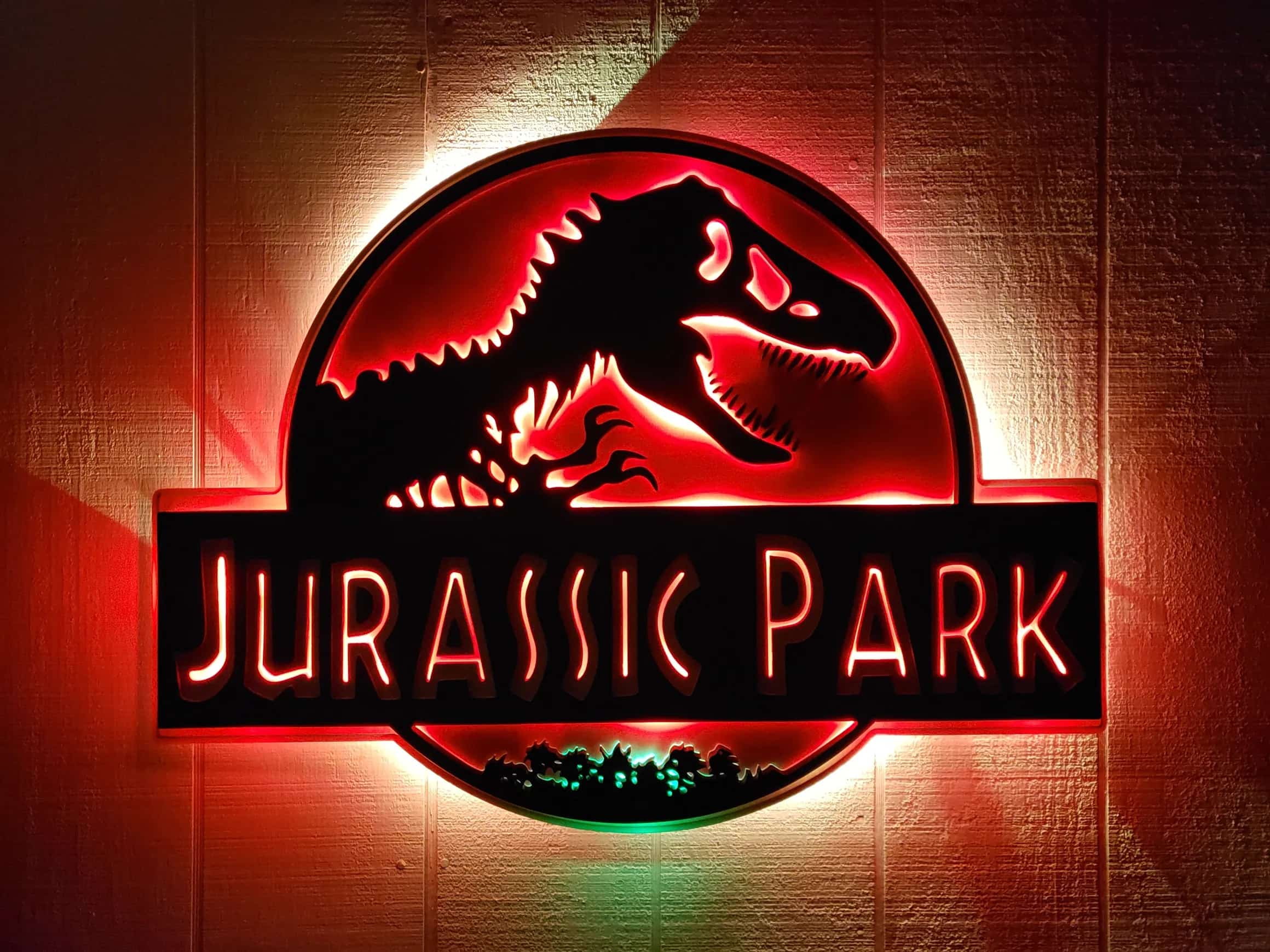 Custom-made, multi-layered Jurassic Park logo sign, depicting the iconic T-Rex silhouette and title. The main body glows with a bright red LED light, while the small ground details at the bottom are illuminated in green, creating a classic, dramatic effect.