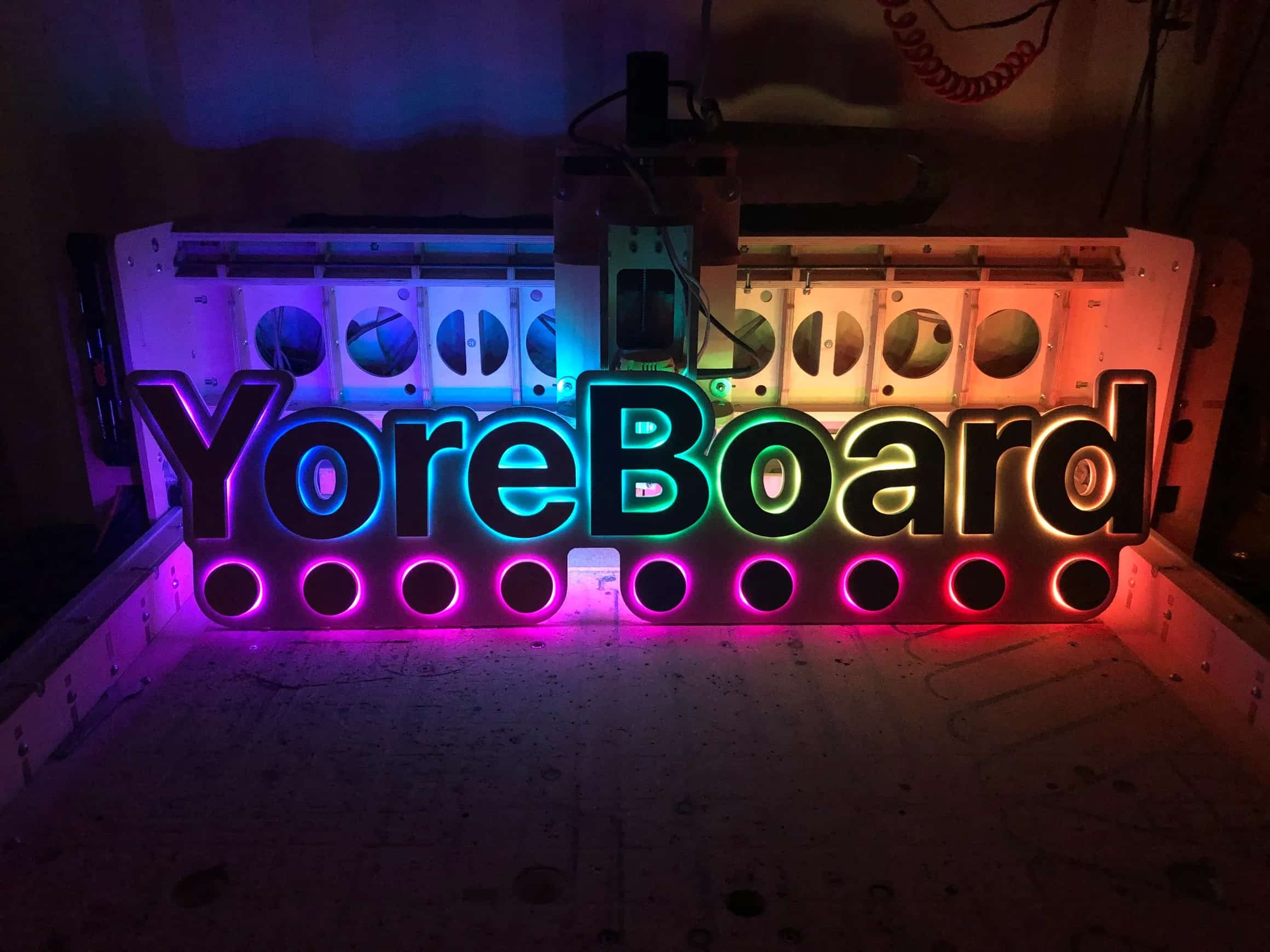Handcrafted YoreBoard brand sign: a multi-layered wooden display with the company name and circular accents, brilliantly lit by a gradient of changing LED colors, demonstrating its visual effects in a dimly lit setting.